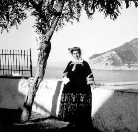 Old black-and-white photograph of a woman in traditional dress standing at a wall above the sea